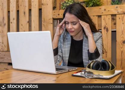 Tired young female remote worker suffering from headache while working on netbook with clipboard and wireless headphones in cafe. Exhausted woman working on laptop in cafe