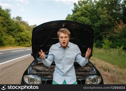 Tired young man at the opened hood, car breakdown. Broken automobile or repairing of vehicle, trouble with auto engine on highway. Tired young man at the opened hood, car breakdown