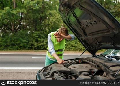 Tired young man at the opened hood fix problem with engine, car breakdown. Broken automobile or repairing of vehicle, trouble with auto on highway