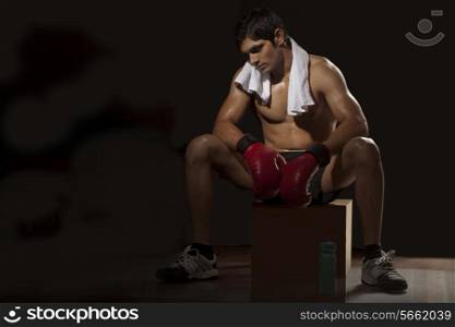 Tired young man wearing boxing gloves resting on stool at gym
