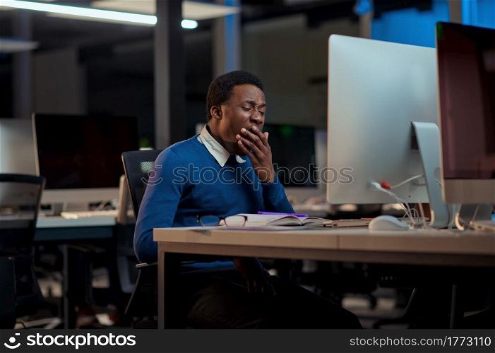 Tired young man works in night office. Sleepy male businessman, dark business center interior on background, modern workplace. Tired young man works in night office