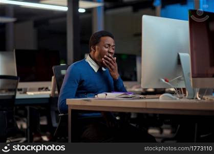 Tired young man works in night office. Sleepy male businessman, dark business center interior on background, modern workplace. Tired young man works in night office