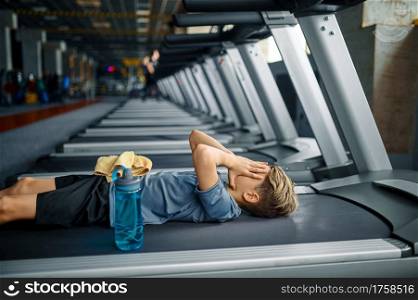 Tired youngster lying on treadmill in gym, running machine. Boy on training in sport club, health care and healthy lifestyle, schoolboy on workout. Tired youngster lying on treadmill in gym