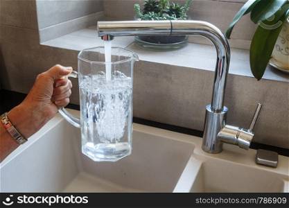 To fill a glass jug with tap water at the sink in a kitchen