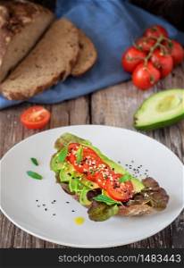 Toast with freshly baked organic bread, olive oil, lettuce, avocado, sesame seeds and cherry tomatoes on a white plate on a dark wooden background. Close-up, lunch concept