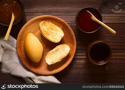 Toasted bread rolls on wooden plate with strawberry and peach jam in rustic bowls and tea on the side, photographed overhead on dark wood with natural light. Bread Rolls with Jam and Tea