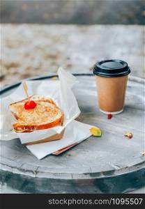 Toasted panini with ham and cheese served on sandwich paper with coffee cup on a wooden outdoor table. Street market food take away concept