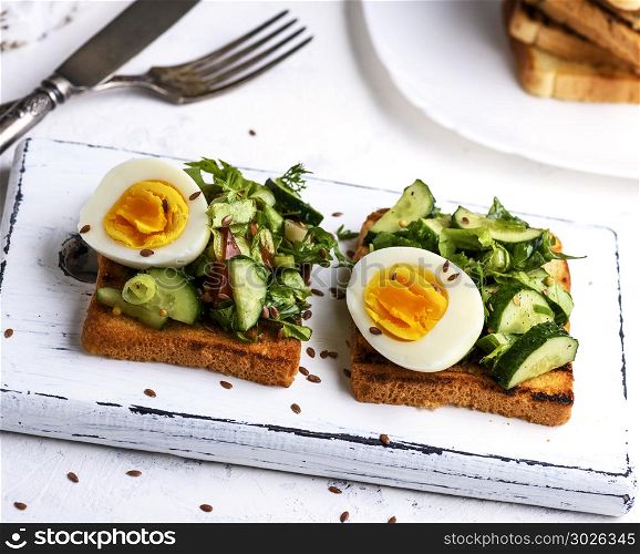 toasted square pieces of bread from white wheat flour with boiled egg, cucumber and green spinach leaves on a white wooden board, breakfast sandwich. pieces of bread from white wheat flour with boiled egg