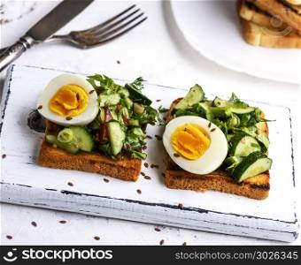 toasted square pieces of bread from white wheat flour with boiled egg, cucumber and green spinach leaves on a white wooden board, breakfast sandwich. pieces of bread from white wheat flour with boiled egg