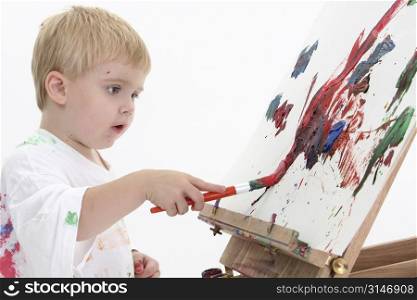 Toddler boy in big white shirt covered in paint at easel. American blonde caucasian boy. Shot over white background.