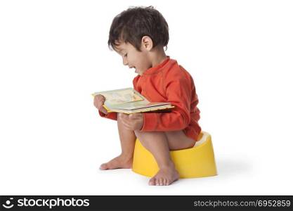 Toddler boy sitting on a potty and reading a book on white background
