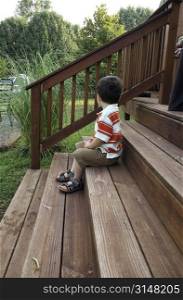 Toddler boy sitting on wooden deck steps early in the morning. Looking away.