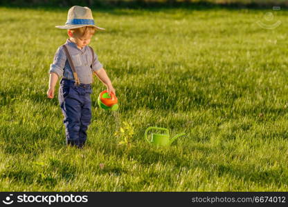 Toddler child outdoors. One year old baby boy wearing straw hat using watering can. Portrait of toddler child outdoors. Rural scene with one year old baby boy wearing straw hat using watering can