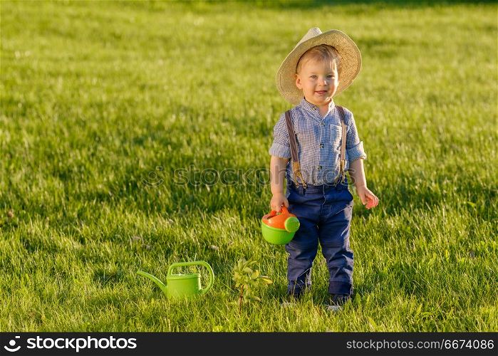 Toddler child outdoors. One year old baby boy wearing straw hat using watering can. Portrait of toddler child outdoors. Rural scene with one year old baby boy wearing straw hat using watering can