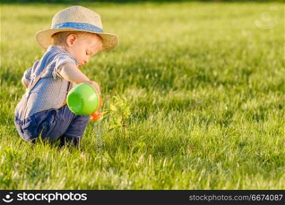 Toddler child outdoors. One year old baby boy wearing straw hat using watering can. Portrait of toddler child outdoors. Rural scene with one year old baby boy wearing straw hat using watering can