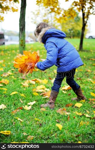 Toddler girl in blue coat picking up leaves on a meadow