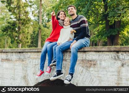 Togetherness and family concept. Friendly father, mother and daughter sit on stone bridge, look from the top, notice beautiful rainbow on sky, indicate on it with fingers, enjoy nature landscapes