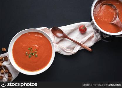 Tomato cream soup in bowl and pot on a black tabletop. Above view of dinner table with vegetarian healthy soup. Flat lay of homemade vegetable soup.