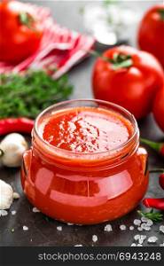Tomato paste, puree in glass jar and fresh tomatos on dark background. Hot vegetable sauce with chili pepper ant tomatoes