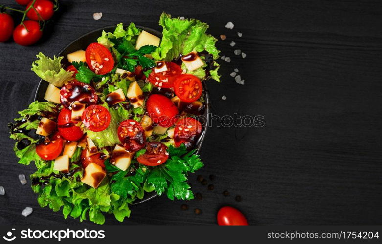 Tomato salad with lettuce, goat cheese, fresh vegetables with sesame seeds and olive oil on a black wooden table. Top view with copy space. Healthy vegetarian food, banner