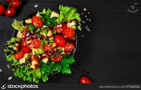 Tomato salad with lettuce, goat cheese, fresh vegetables with sesame seeds and olive oil on a black wooden table. Top view with copy space. Healthy vegetarian food, banner