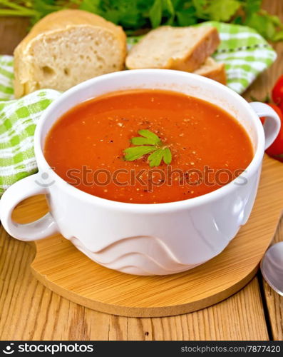 Tomato soup in a white bowl, napkin, spoon, tomato, parsley, bread on a wooden boards background