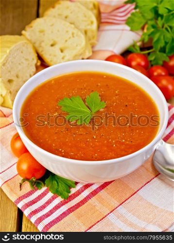 Tomato soup in a white bowl on a napkin with a spoon, tomato, parsley, bread on a wooden boards background