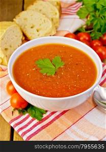 Tomato soup in a white bowl on a napkin with a spoon, tomato, parsley, bread on a wooden boards background