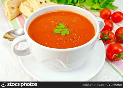 Tomato soup in a white cup and saucer, tomatoes, bread, parsley on the background of striped linen tablecloth