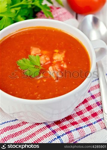 Tomato soup with chunks of vegetable in a white bowl on a napkin with a spoon, tomatoes, parsley on a lighter background board