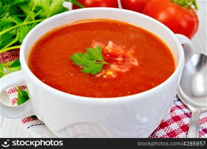 Tomato soup with chunks of vegetable in a white bowl on a napkin with a spoon, tomatoes, parsley on a lighter background board
