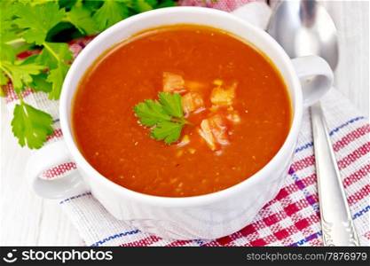 Tomato soup with pieces of vegetables in a white bowl on a napkin, spoon, tomato, parsley on a light background boards