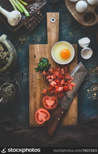 Tomatoes and eggs on wooden cutting board with knife on dark rustic kitchen table background. Shakshuka cooking ingredients. Top view. Healthy food eating