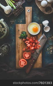 Tomatoes and eggs on wooden cutting board with knife on dark rustic kitchen table background. Shakshuka cooking ingredients. Top view. Healthy food eating