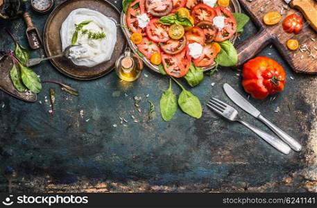 Tomatoes and mozzarella salad, preparation on dark aged rustic background, top view. Italian lunch with tomatoes and mozzarella, cutlery and cooking ingredients. Italian food concept