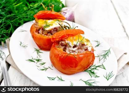 Tomatoes stuffed with meat and rice with cheese in a plate on a napkin, fork, dill and parsley on a wooden board background