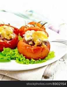 Tomatoes stuffed with meat and rice with cheese on lettuce in a plate on a napkin, fork against a light wooden board