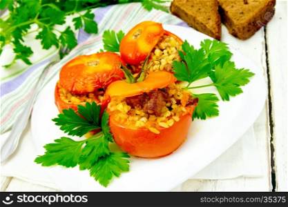 Tomatoes stuffed with meat and steamed wheat bulgur in a white plate, napkin, fork, bread and parsley on the background light wooden boards