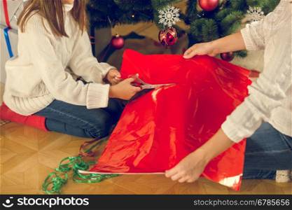 Toned closeup image of girl and mother sitting under Christmas tree and cutting red wrapping paper for presents