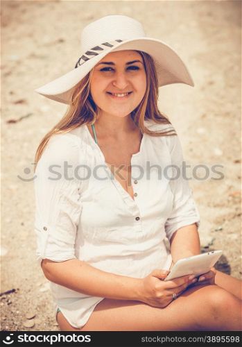 Toned closeup portrait of smiling woman in hat using tablet on beach