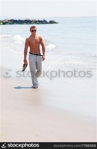 Toned cute handsome male walking on the beach with naked torso showing six pack abs holding shoes in hand wearing sunglasses