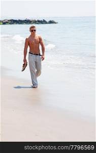 Toned cute handsome male walking on the beach with naked torso showing six pack abs holding shoes in hand wearing sunglasses