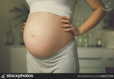 Toned image of pregnant woman posing at window and holding hands on belly