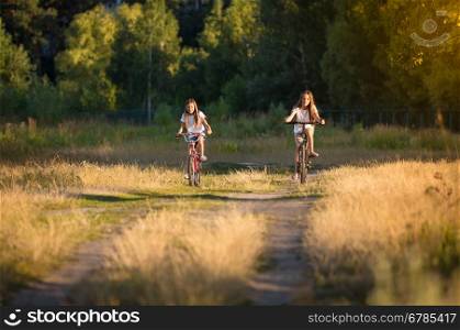 Toned image of two teenage girls riding bicycles on meadow at sunset