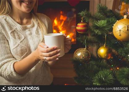 Toned image of woman in woolen sweater warming at fireplace with cup of tea