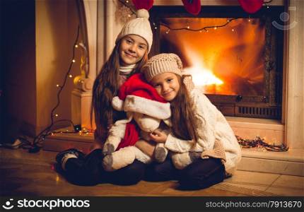 Toned old styled photo of two happy girls sitting with toy at fireplace on Christmas eve