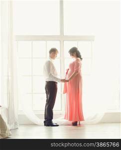Toned photo of beautiful pregnant couple holding hands at big window