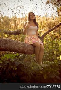 Toned photo of cute young woman sitting on big branch at foreast