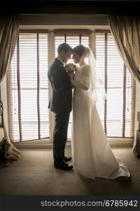 Toned photo of happy bride and groom embracing at big window at hotel room