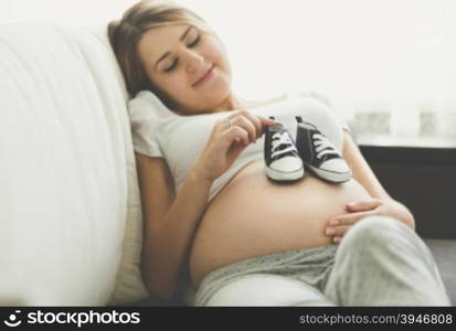 Toned photo of happy pregnant woman lying on sofa and holding baby boots